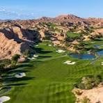 Golf course nestled in desert landscape with green fairways, sand traps, and brown rocky hills under blue sky