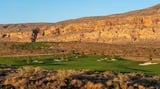 Desert golf course with green fairways nestled against towering sandstone cliffs under blue sky